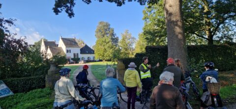 Groep fietsers luistert naar een gids bij een landhuis in een groene, bosrijke omgeving.