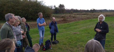 Groep mensen luistert naar een persoon die buiten op een grasveld spreekt, omringd door bomen.