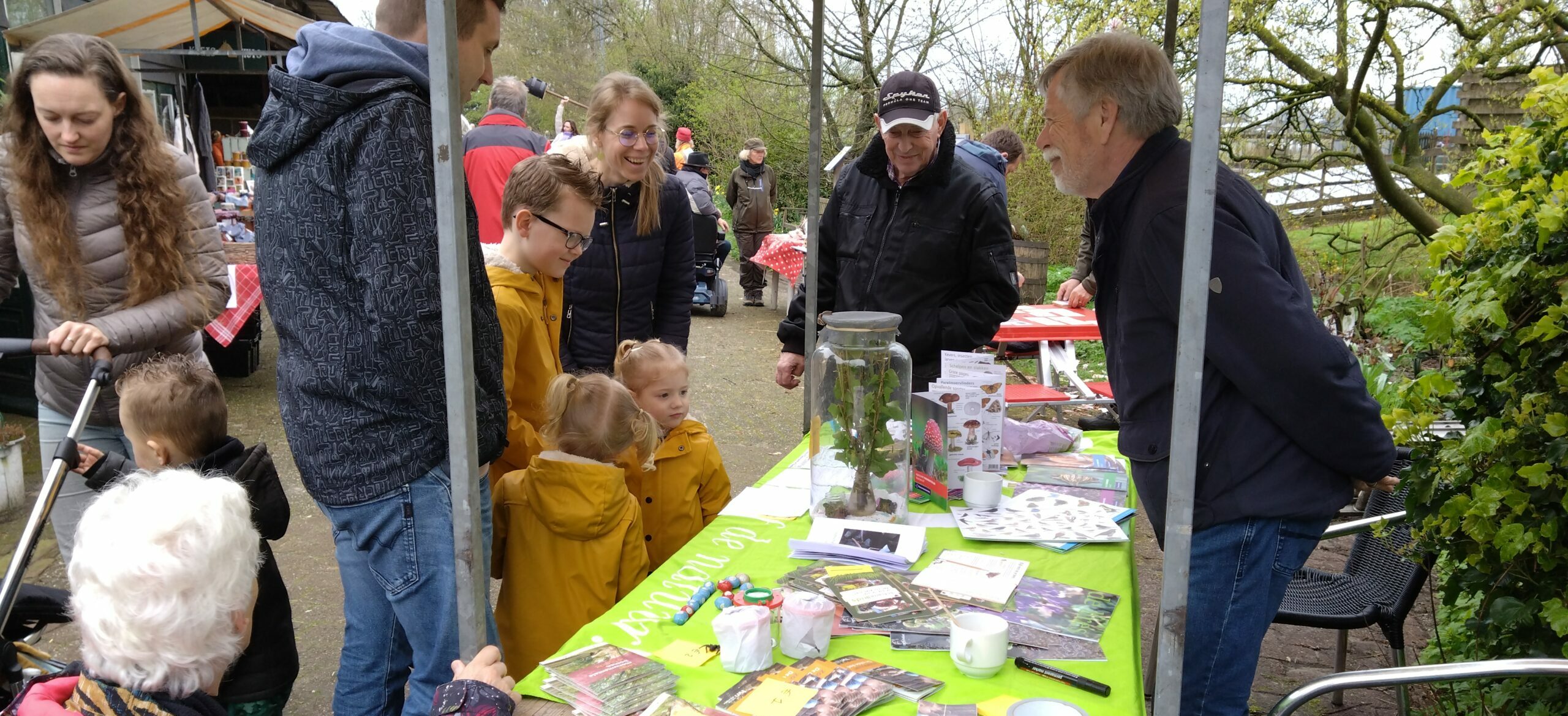 Mensen bij een kraam op een buitenmarkt, met brochures en items op een groene tafel.