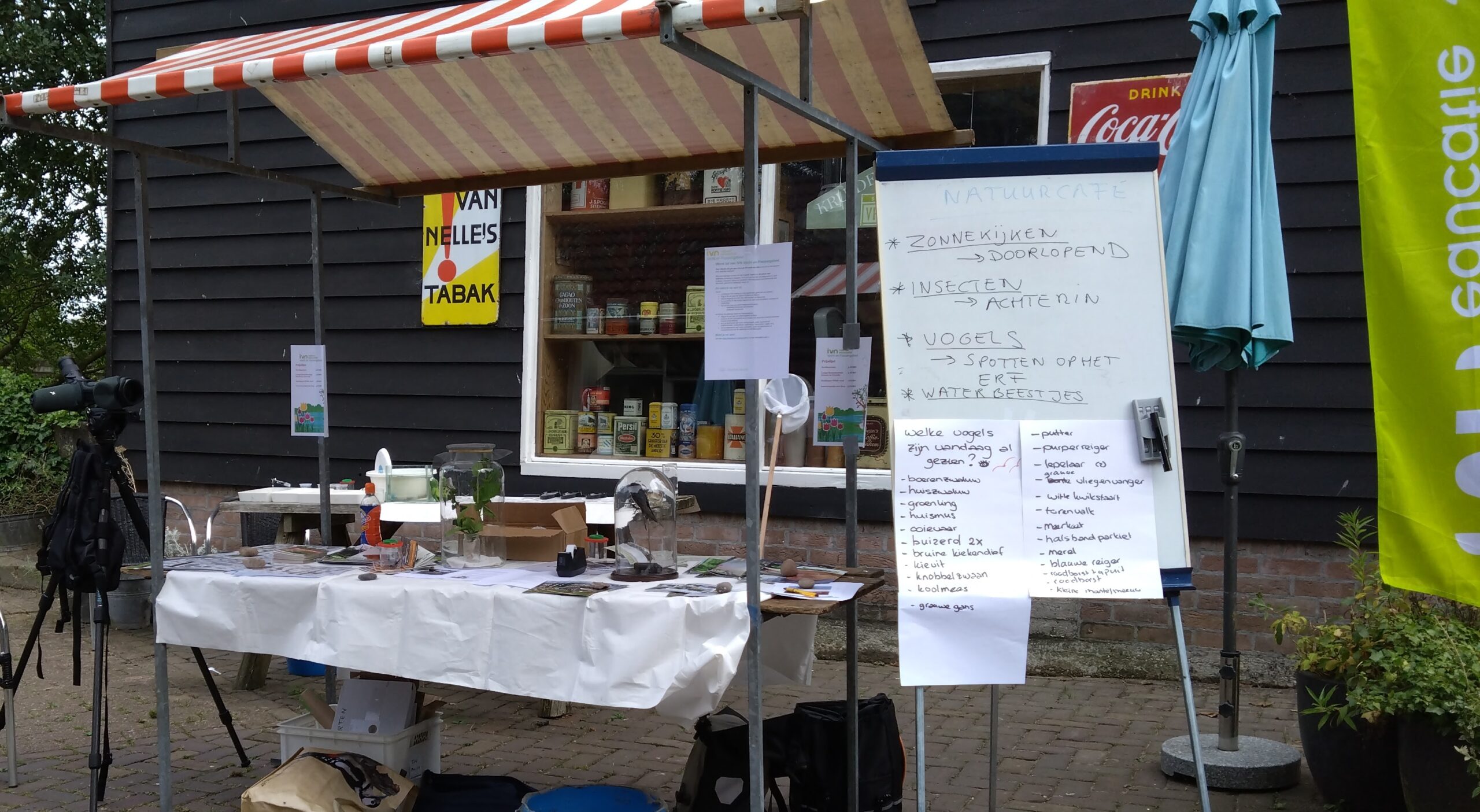 Tafel met natuurbenodigdheden, telescoop, en whiteboard met tekst bij zwarte houten schuur.
