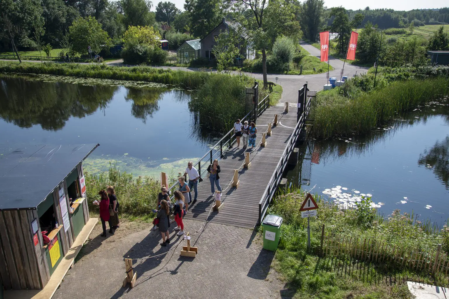 Mensen steken een brug over bij een vijver in een groene omgeving met duidelijk zonlicht.