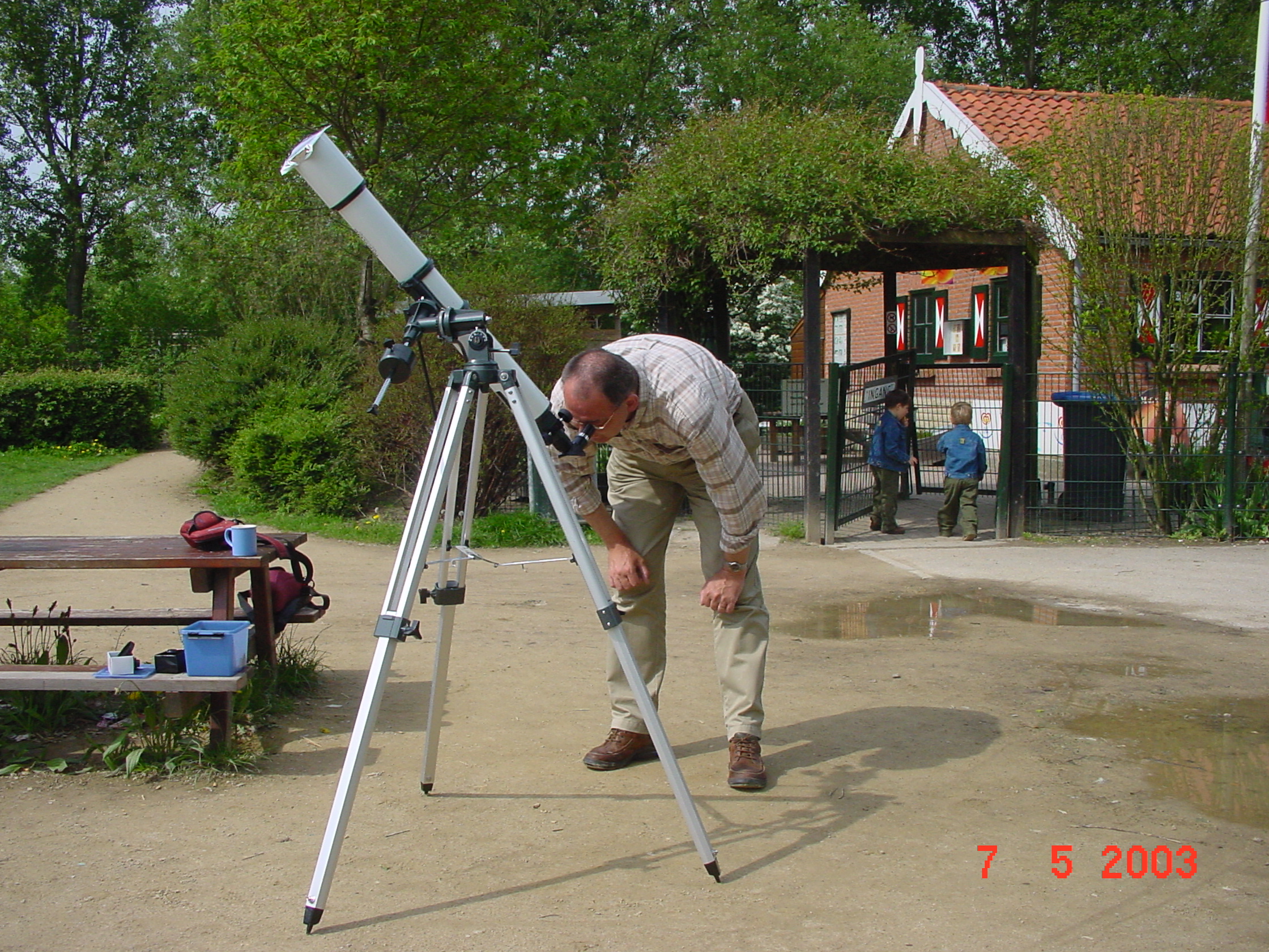 Man kijkt door telescoop in park, kinderen spelen bij poort; datum: 7 mei 2003.