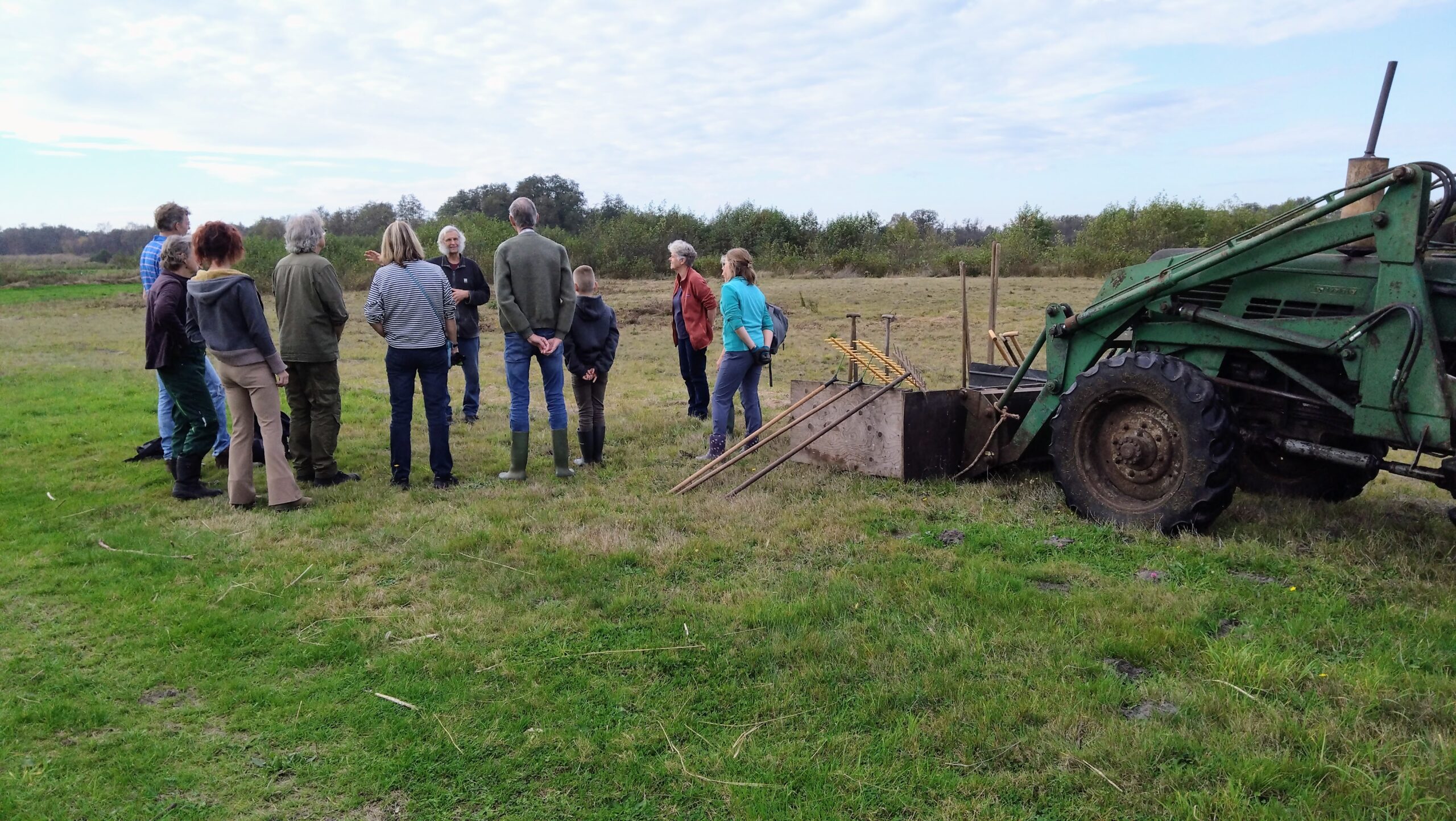 Groep mensen luistert buiten naast een groene tractor op een veld met gras en gereedschap.
