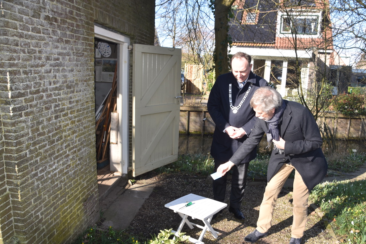 Twee mannen bij een open schuurdeur, een legt iets op een wit tafeltje in een tuin.
