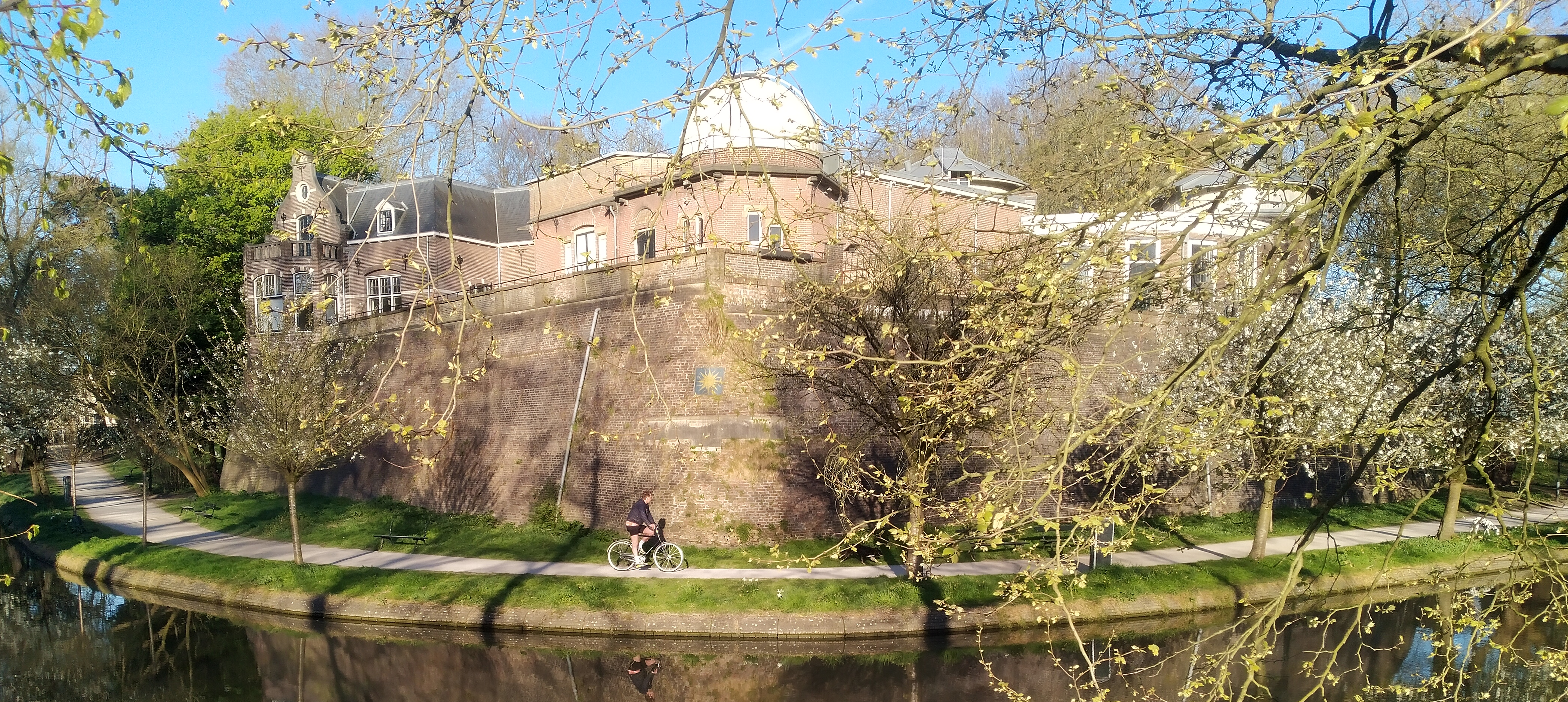 Historisch gebouw met koepel en bakstenen muur, omgeven door bomen en gracht; fietser op pad.