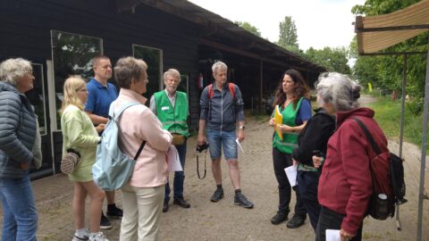 Een groep mensen luistert naar een vrouw in een groen hesje tijdens een buitenbijeenkomst.