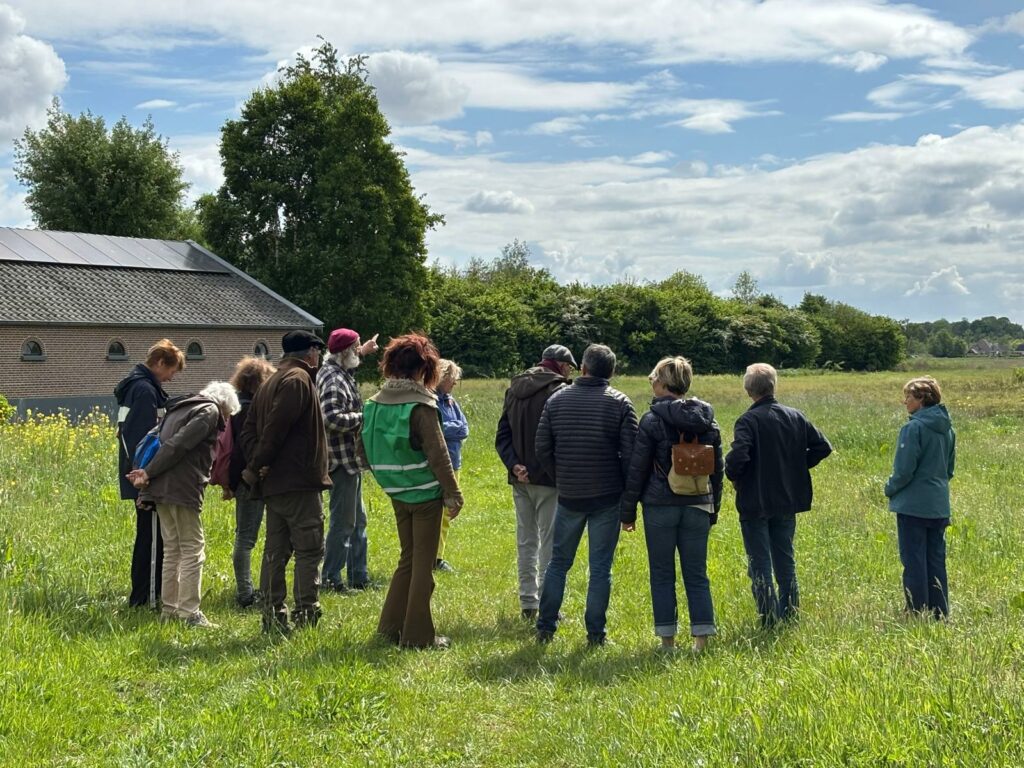 Groep mensen op grasveld naast gebouw met zonnepanelen, kijken naar een gids die wijst naar de natuur.
