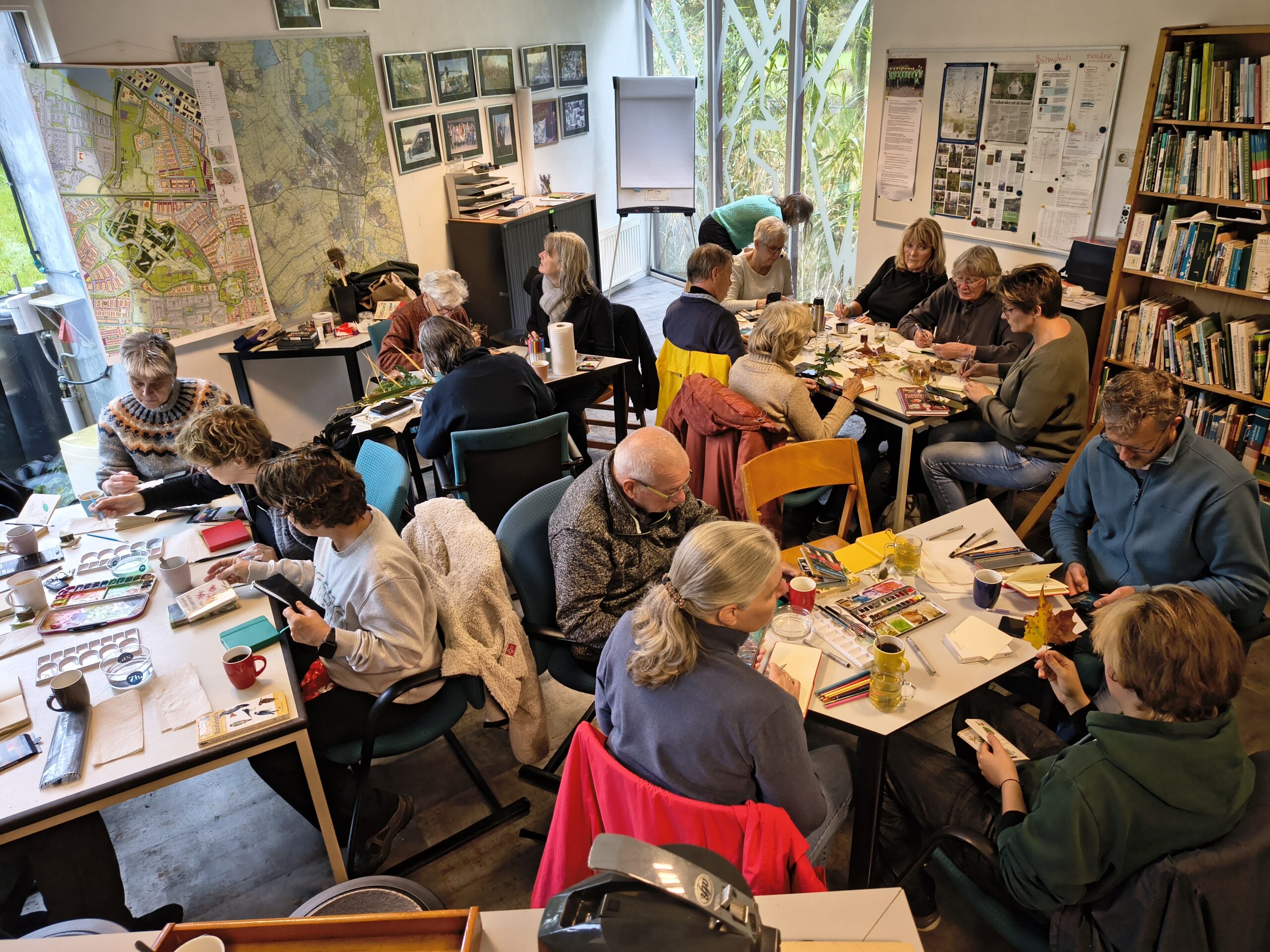 Een groep mensen schildert en tekent aan tafels in een kamer met kaarten en boeken.