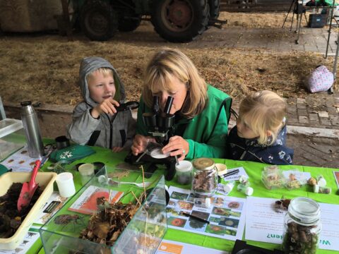 Volwassene en twee kinderen onderzoeken natuurvoorwerpen met een microscoop aan een tafel vol monstermateriaal.