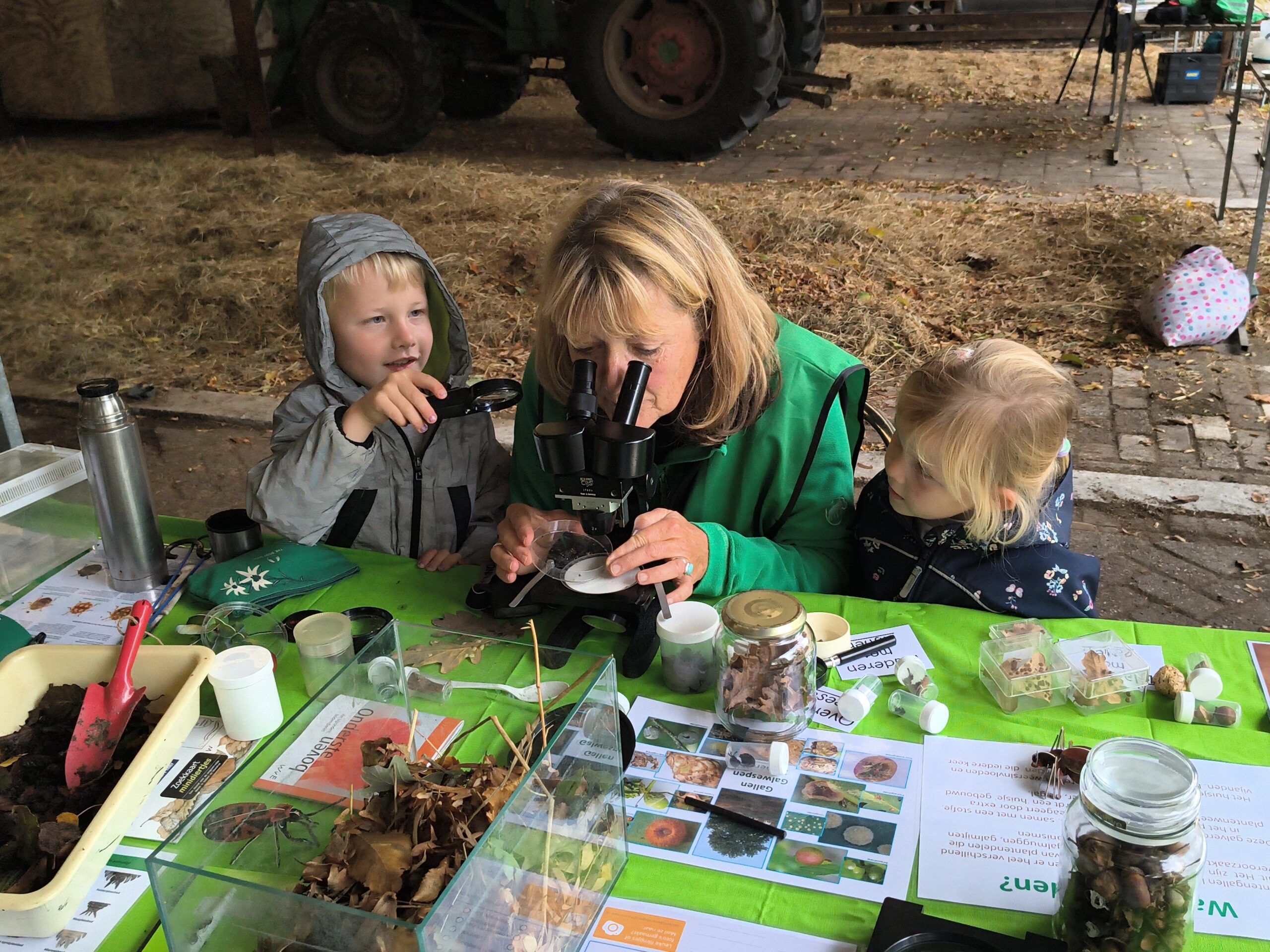 Volwassene en twee kinderen onderzoeken natuurvoorwerpen met een microscoop aan een tafel vol monstermateriaal.