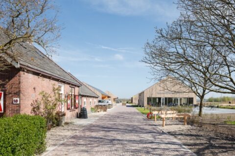 Landelijk pad met stenen boerderij links en modern houten gebouw rechts onder blauwe lucht.