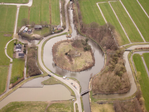 Luchtfoto van groen landschap met wegen en waterwegen rond een bebost eilandje.