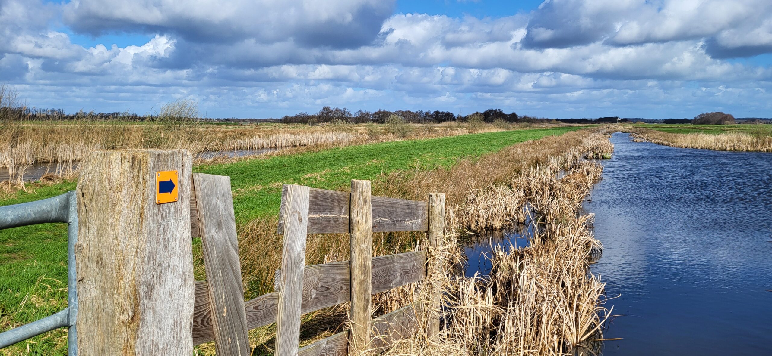 Houten hek met routebordje langs kanaal in groen, open landschap onder wolkenlucht.