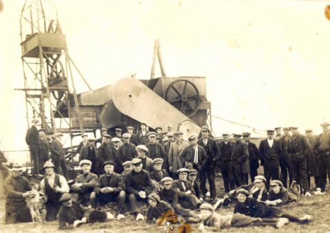 Grote groep mannen poseert voor een industriële machine. Ze dragen petten en werkkleding.