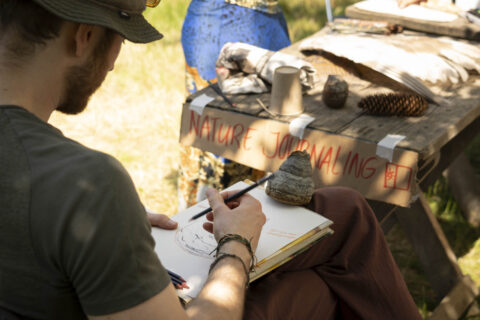 Man schetst natuur in notitieboek bij tafel met bord "Nature Journaling" en natuurvoorwerpen.