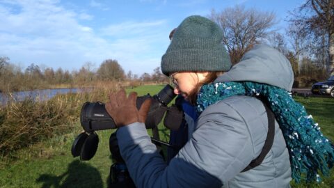 Persoon met warme kleding kijkt door een telescoop bij rivier in winterlandschap.