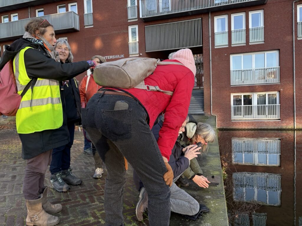Groep mensen observeert water bij moderne bakstenen gebouwen onder zonnige omstandigheden.
