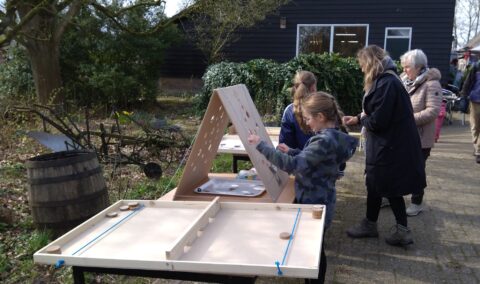 Kinderen spelen een tafelspel buiten, naast een houten ton en oude landbouwapparatuur.