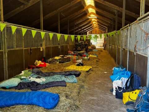 Slaapzakken en rugzakken op stro in een stal, versierd met groene vlaggetjes.