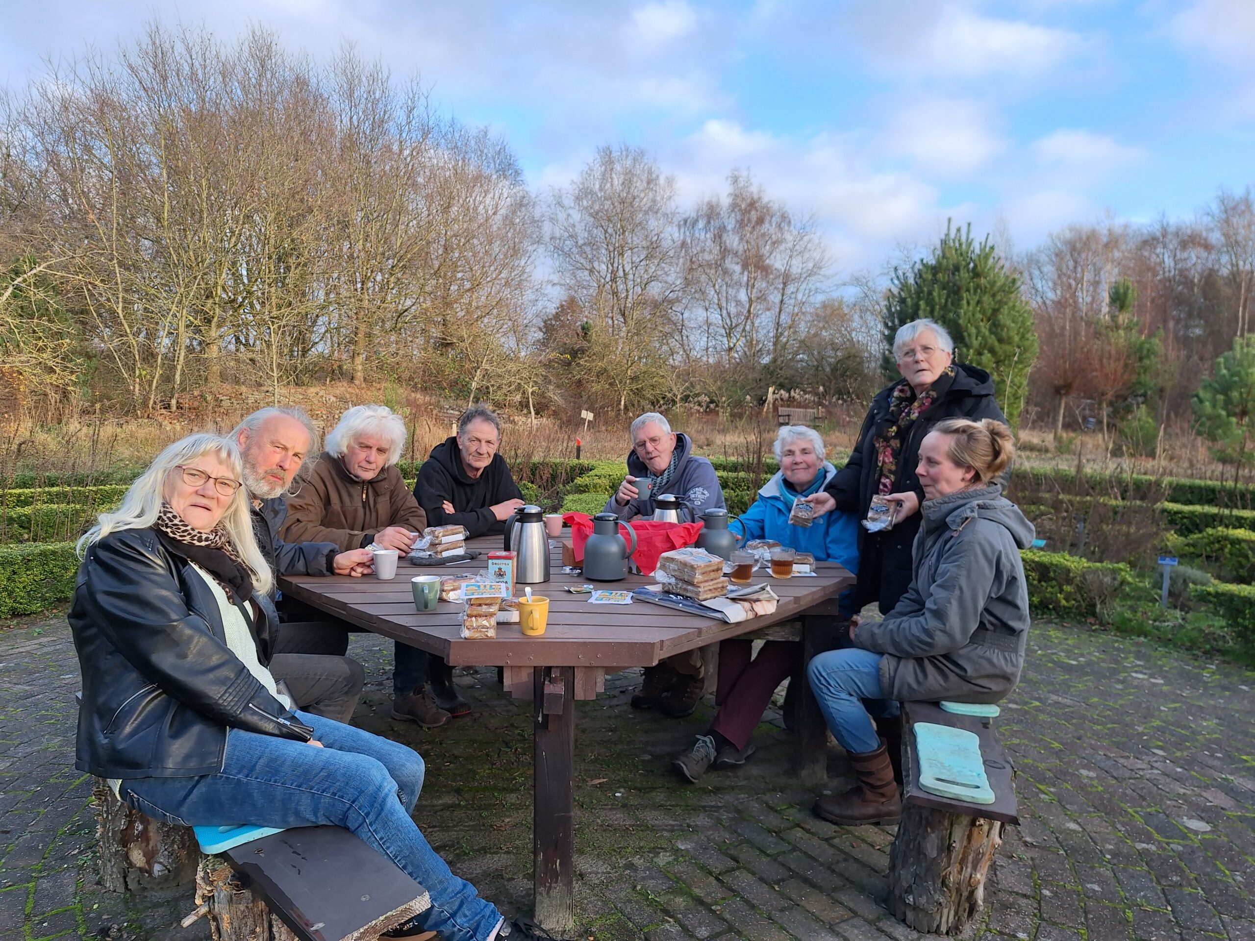 Groep mensen zit aan picknicktafel buiten, met drankjes en snacks, omgeven door bomen.