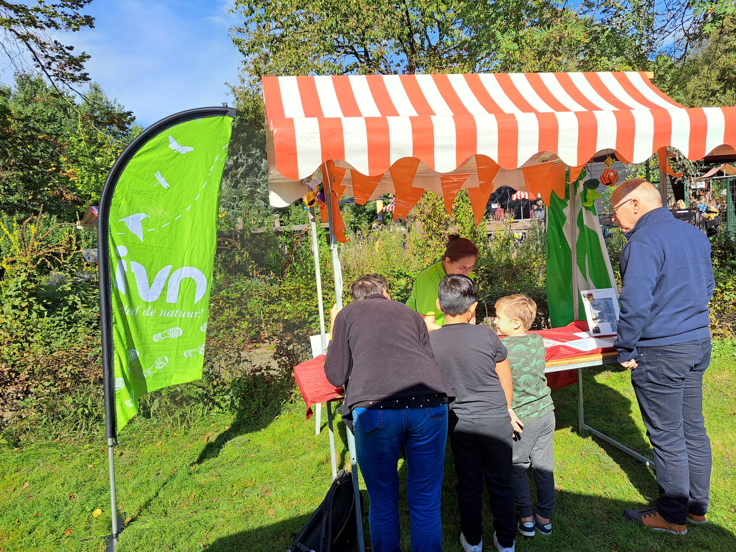 Mensen bij een informatiestand met rood-wit gestreepte luifel, groene vlag met tekst "IVN".