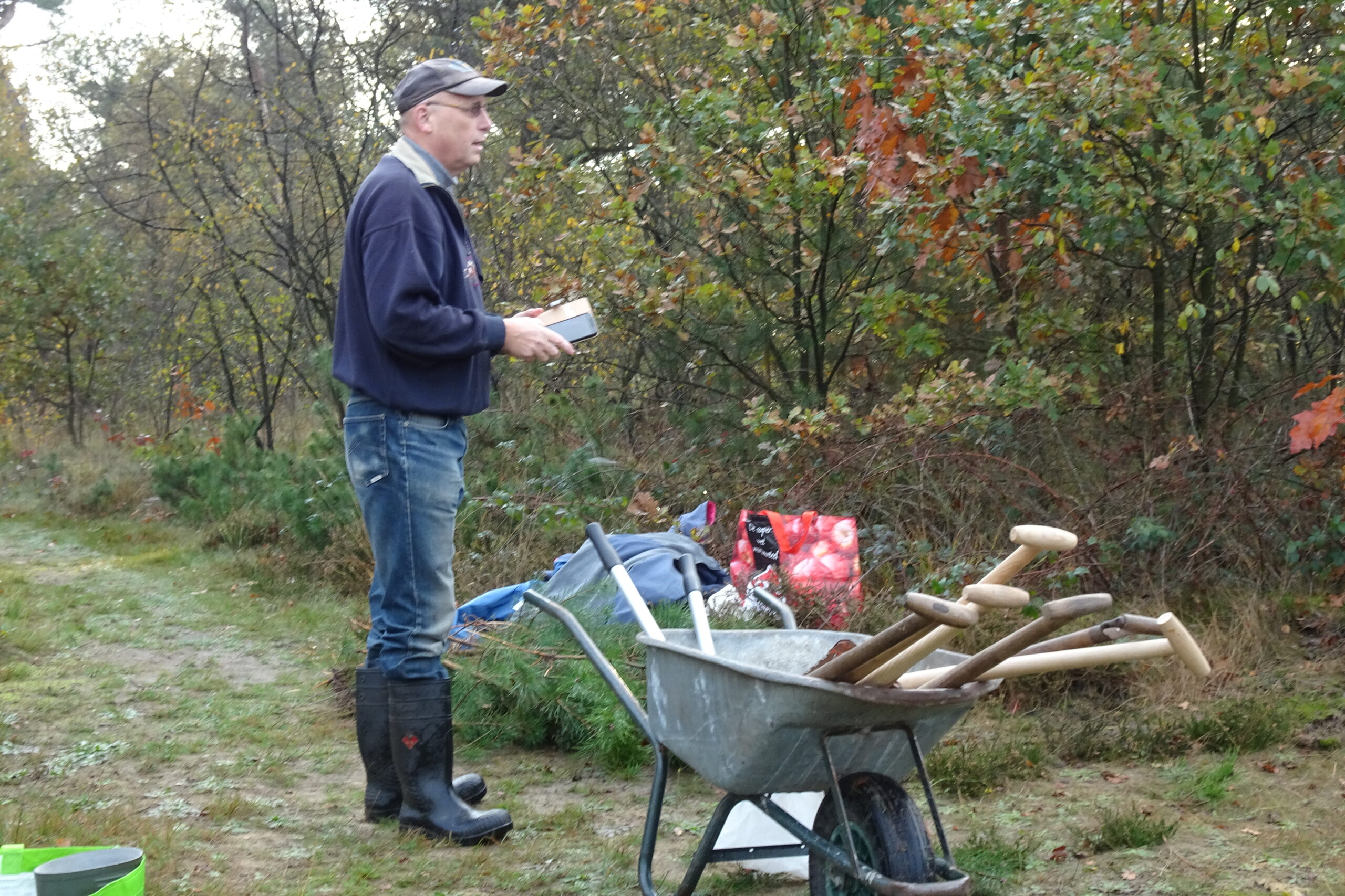Man met pet bij kruiwagen vol schoppen in bosrijke omgeving.