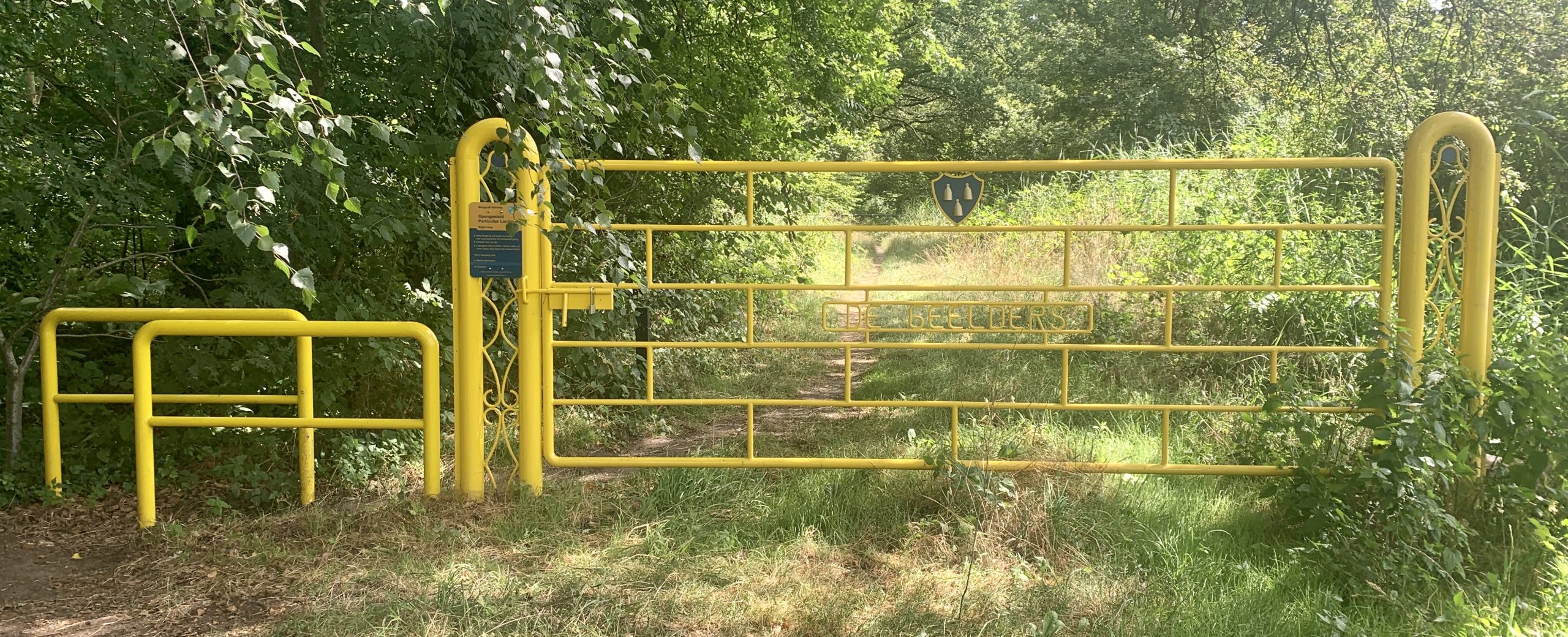 Gele toegangspoort in een bos met een wandelpad dat verder leidt. Op de poort staat "De Geelders".