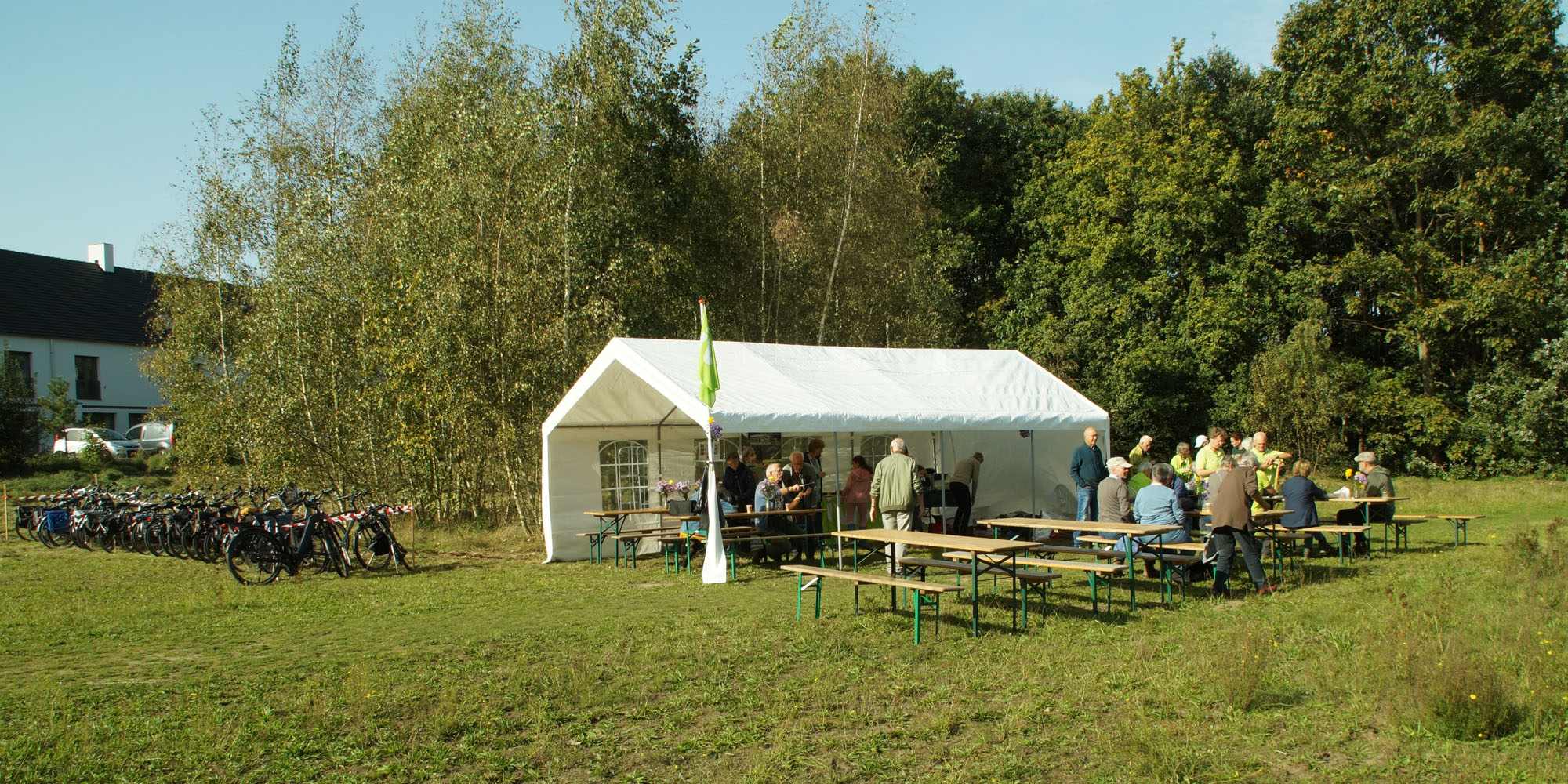 Een groep mensen zit bij een witte tent in een groene open ruimte met fietsen rechts.