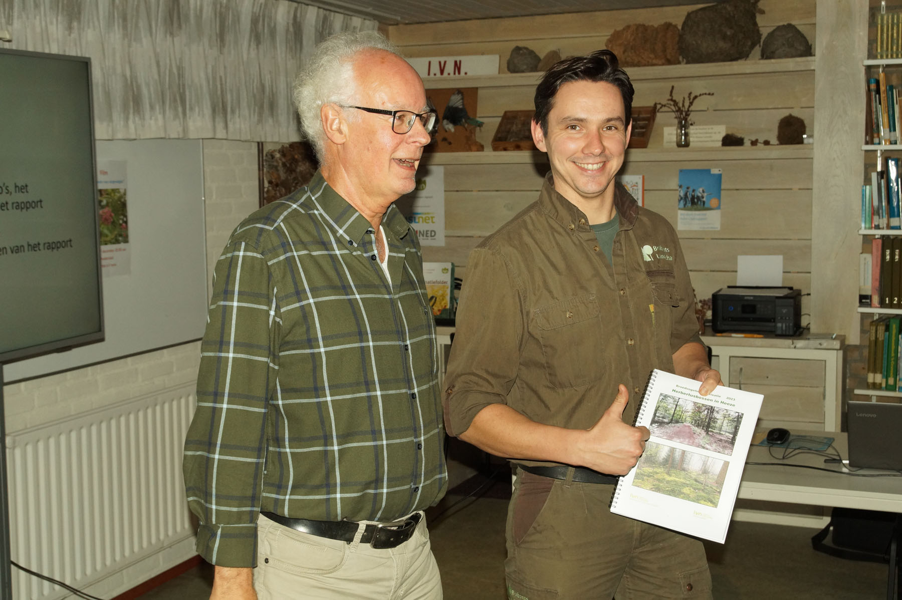 Twee mannen in een kamer; een houdt een rapport vast en geeft een duim omhoog.