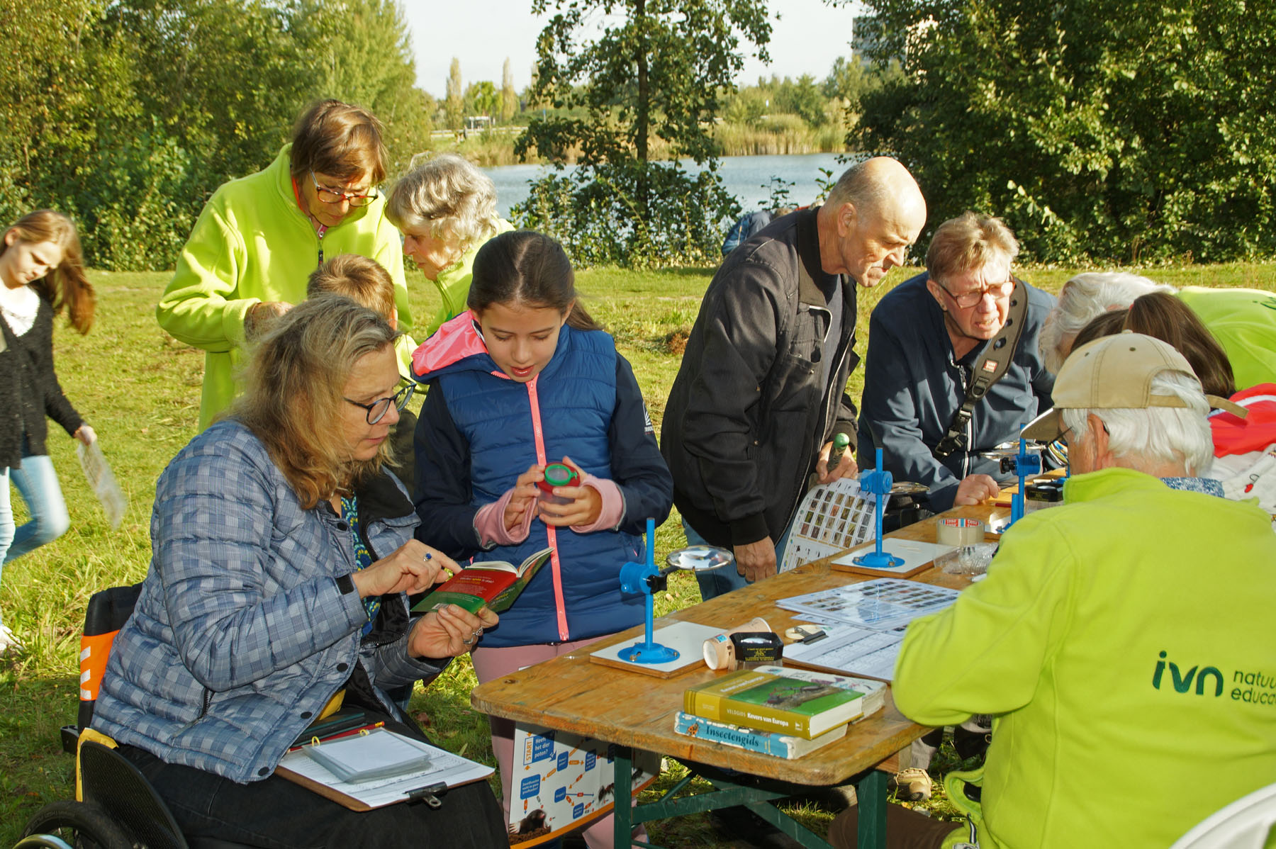 Groep mensen onderzoekt natuurboeken en items aan een tafel in een parklandschap.