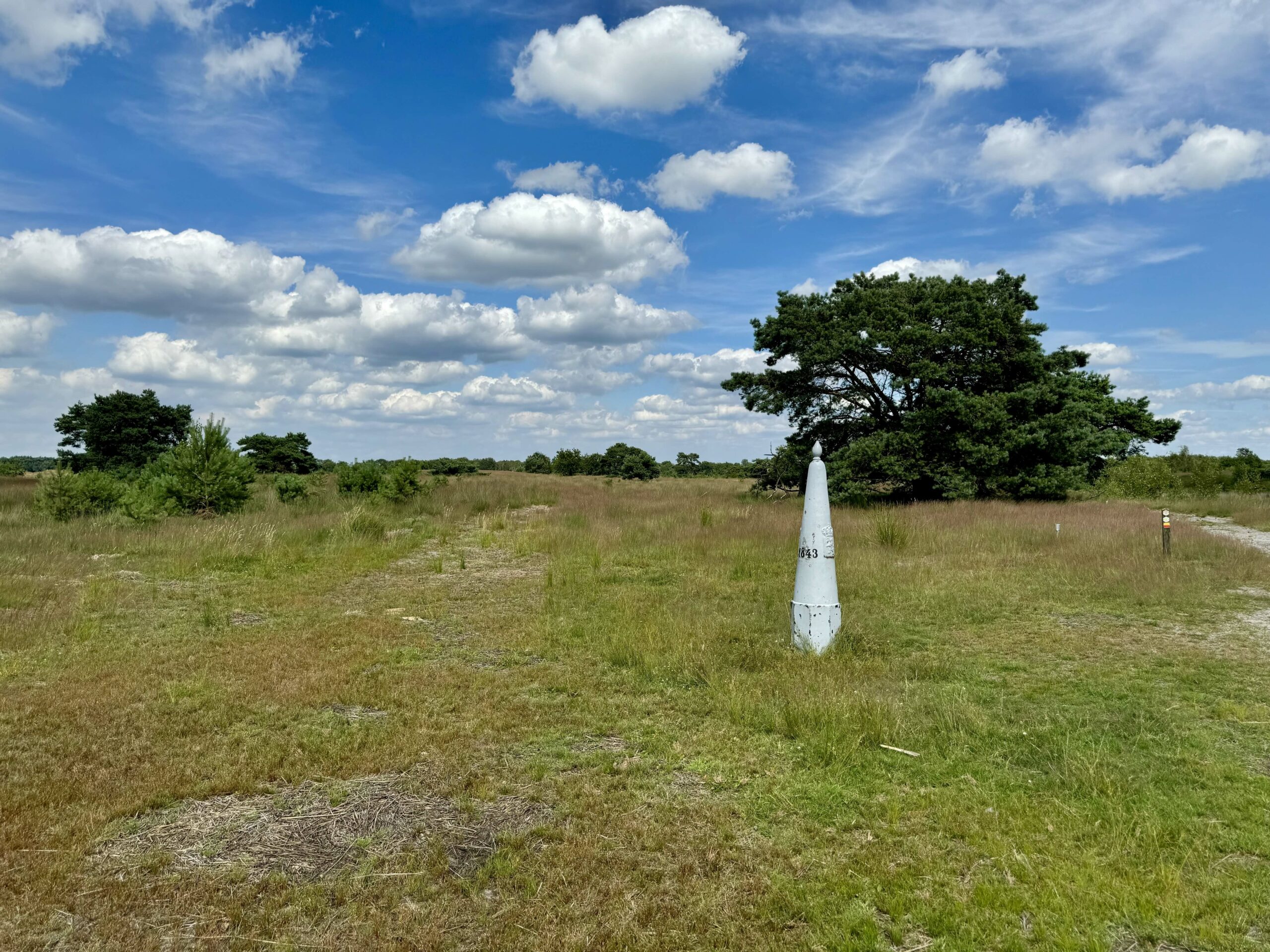 Grasveld met een grenspaal en enkele bomen onder een blauwe lucht met wolken.