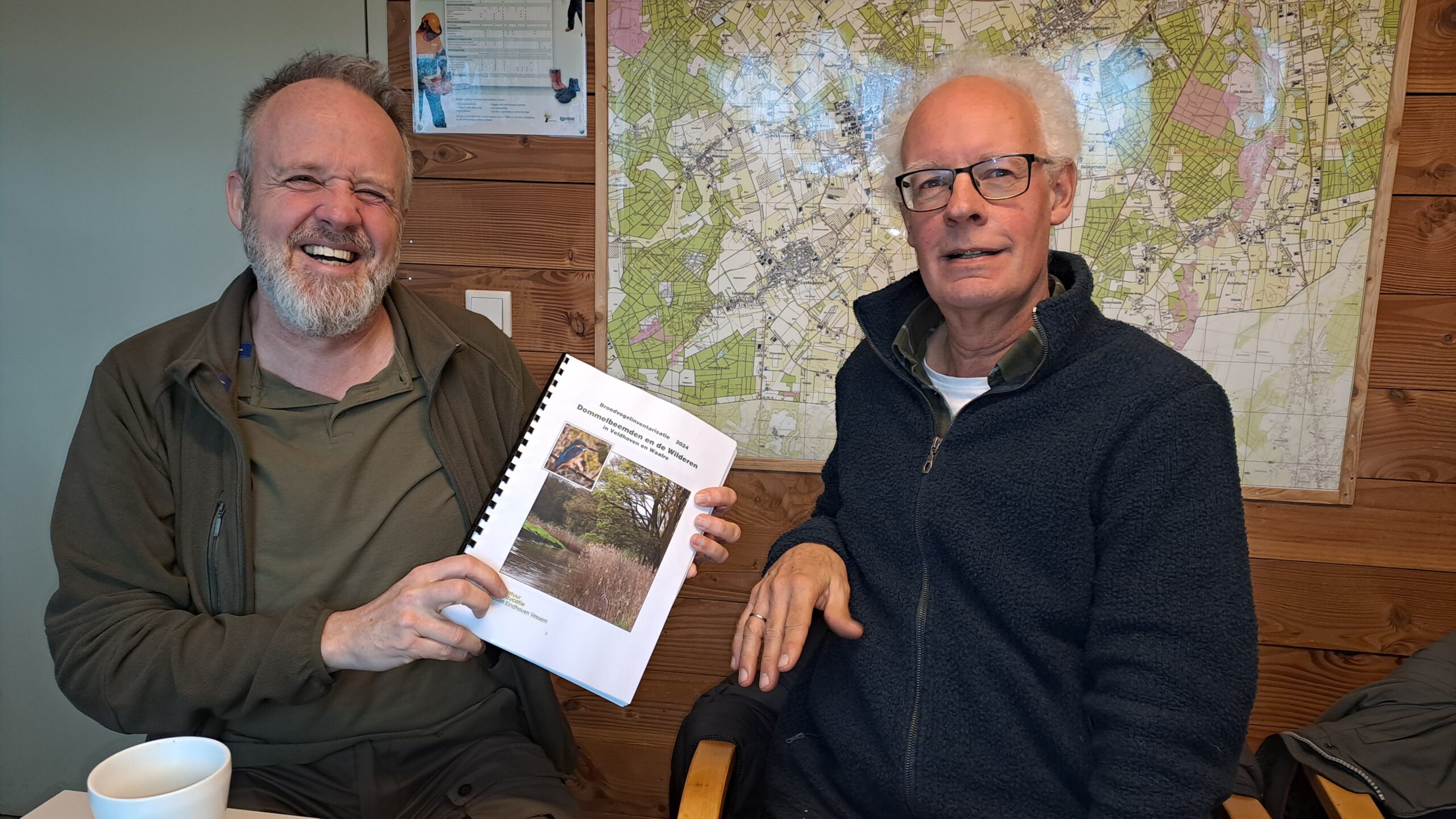 Twee mannen met een rapport voor een kaart aan de muur. De linker man glimlacht breed.