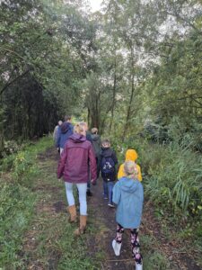 Groep wandelt op bosachtig pad onder groene bomen. Diverse regenjassen en rugzakken zichtbaar.