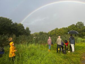Een groep mensen in een veld met een regenboog en paraplu, kind in gele jas met schepnet.