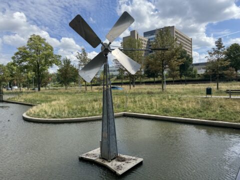 Windturbine in een vijver, omgeven door gras en bomen, met een gebouw op de achtergrond.