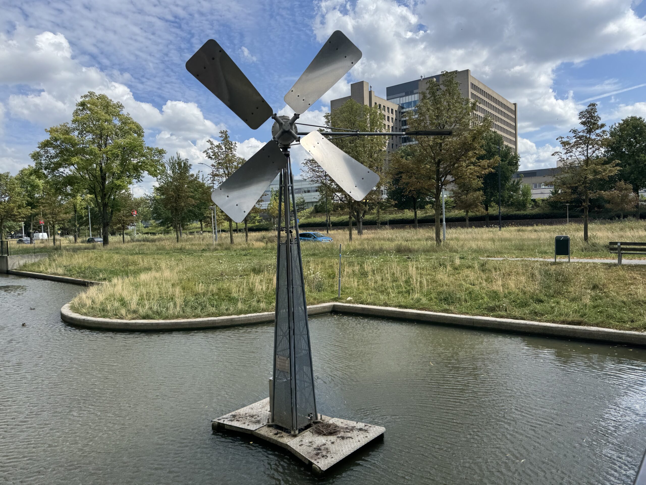Windturbine in een vijver, omgeven door gras en bomen, met een gebouw op de achtergrond.