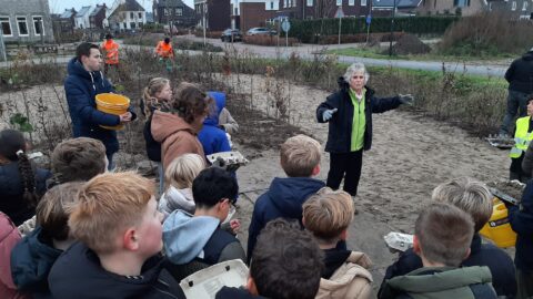 Groep kinderen luistert naar een instructeur in een buitenomgeving met een man die een gele emmer vasthoudt.
