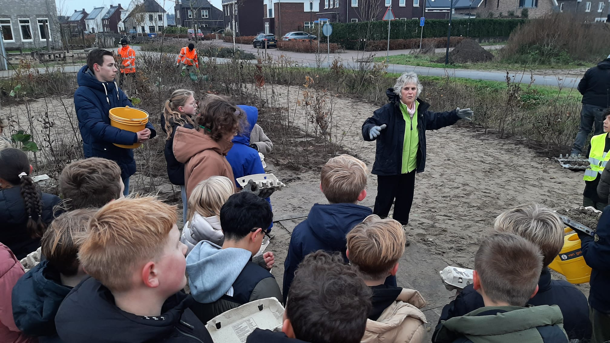 Groep kinderen luistert naar een instructeur in een buitenomgeving met een man die een gele emmer vasthoudt.