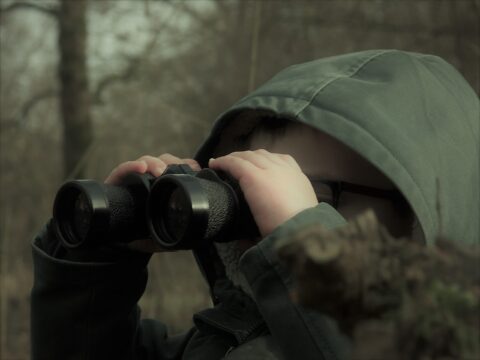 Een kind in een capuchon kijkt door een verrekijker in een bosrijke omgeving.
