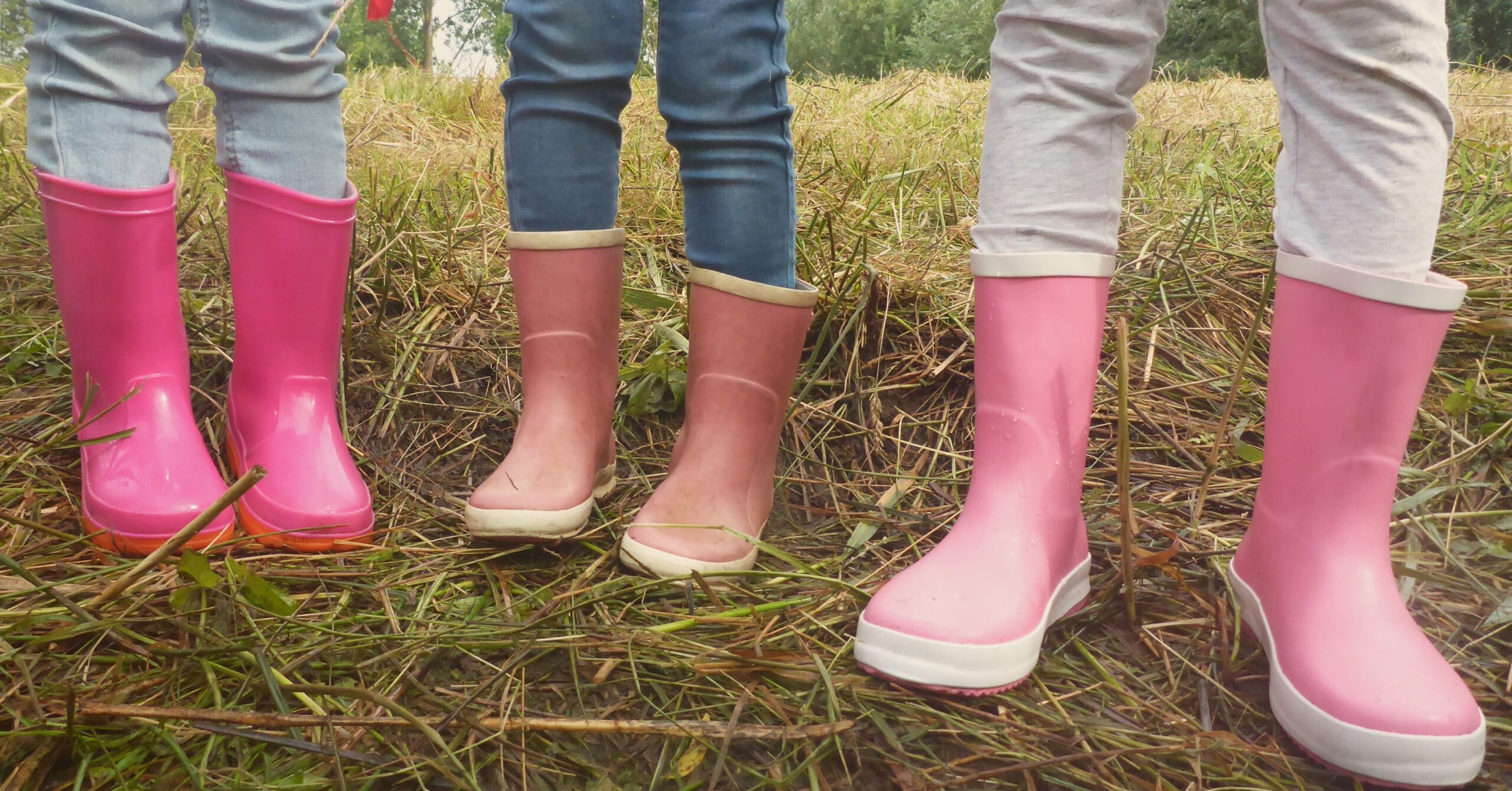 Drie paar benen in regenlaarzen op een modderig veld. Twee roze, één lichtroze.