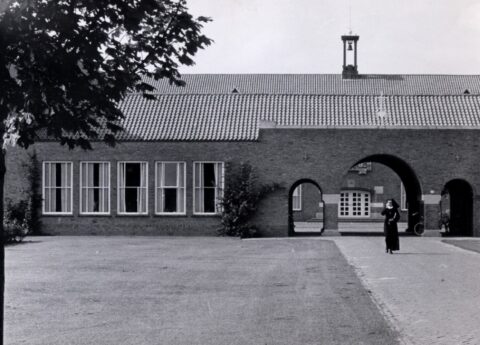 Bakstenen schoolgebouw met bogen, een non loopt op het pad.