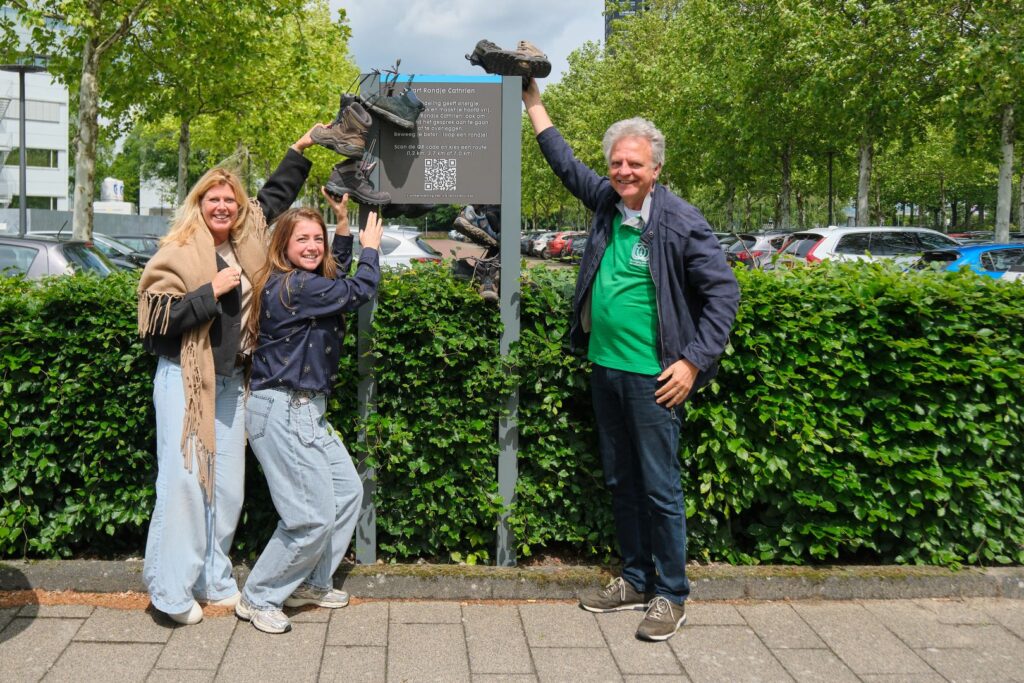 Drie mensen poseren lachend met wandelschoenen bij een bord in groene buitenomgeving.