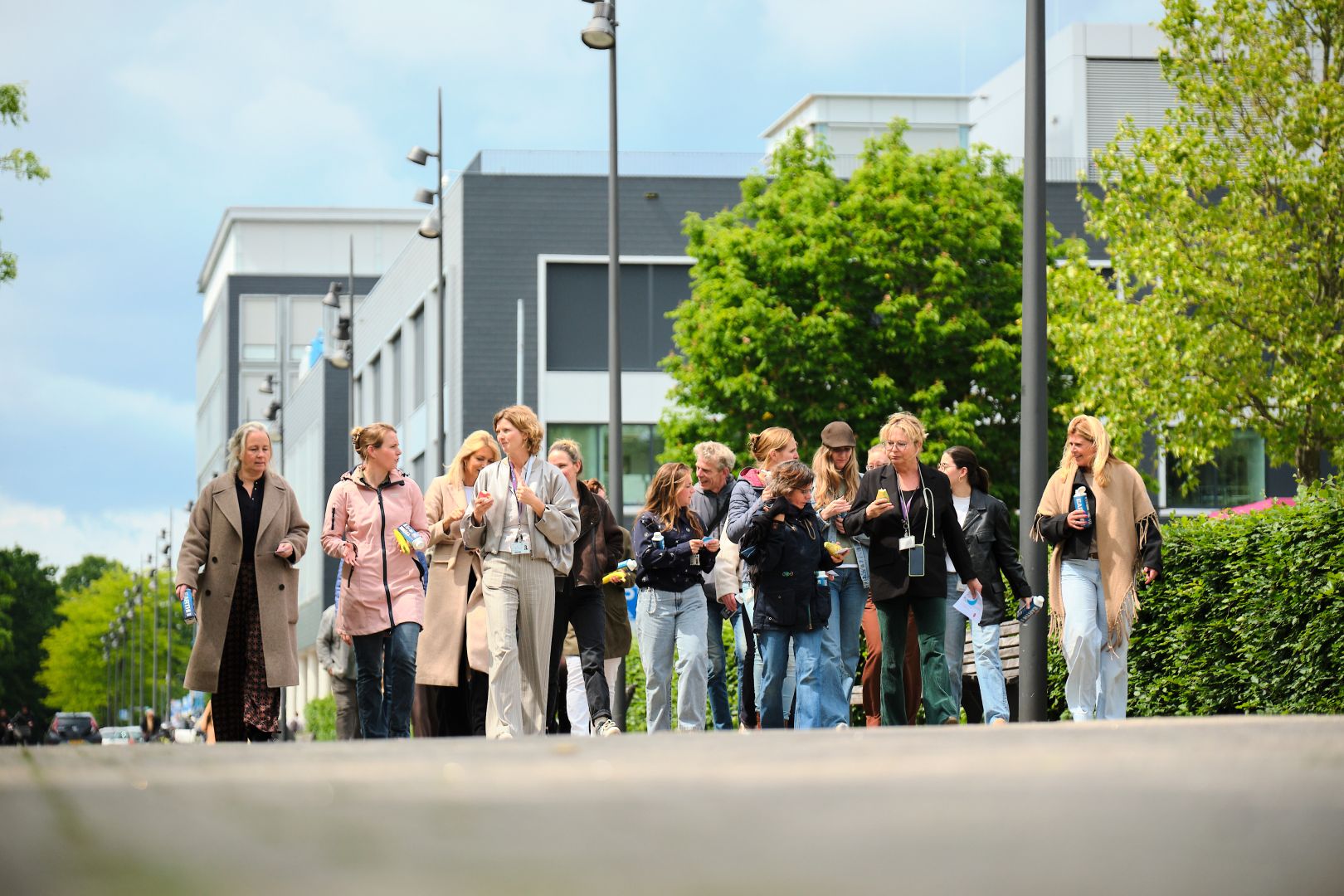 Mensen wandelen samen op een stadsweg naast moderne gebouwen en groen, onder een bewolkte hemel.