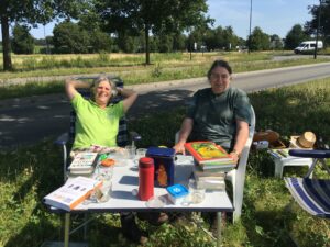 Twee mensen relaxen buiten met boeken en drankjes aan een tafel langs een weg met gras en bomen.