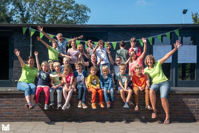 Groep enthousiaste kinderen en volwassenen poseert voor een gebouw, met groene vlaggetjes op de achtergrond.