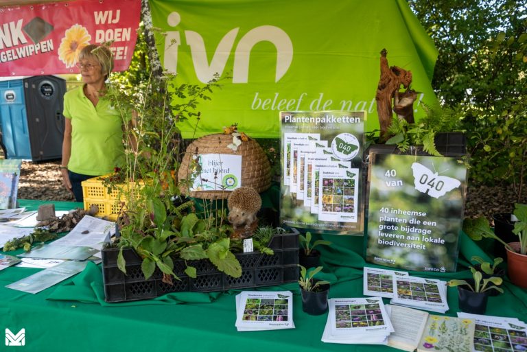 Beursstand met plantenpakketten, promotieposters en een medewerker in groene outfit.