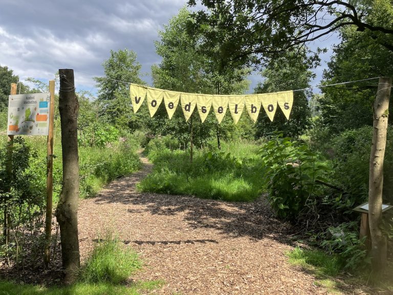 Ingang van een voedselbos met bord en slinger, omringd door groene bomen en een pad.