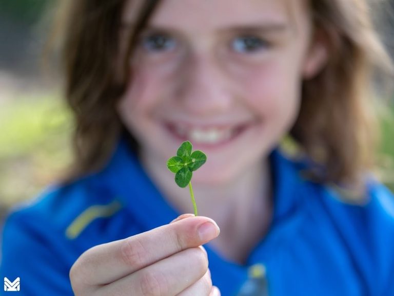 Kind in blauwe jas houdt een klavertjevier vast en glimlacht naar de camera.