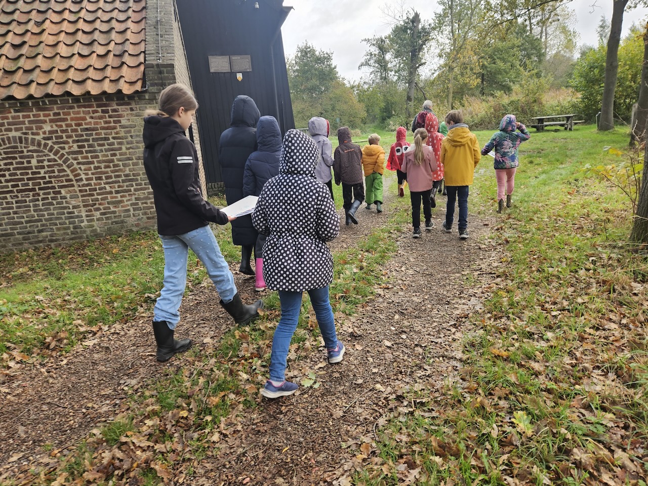 Kinderen wandelen in herfstkleding langs een bospad bij een bakstenen gebouw.