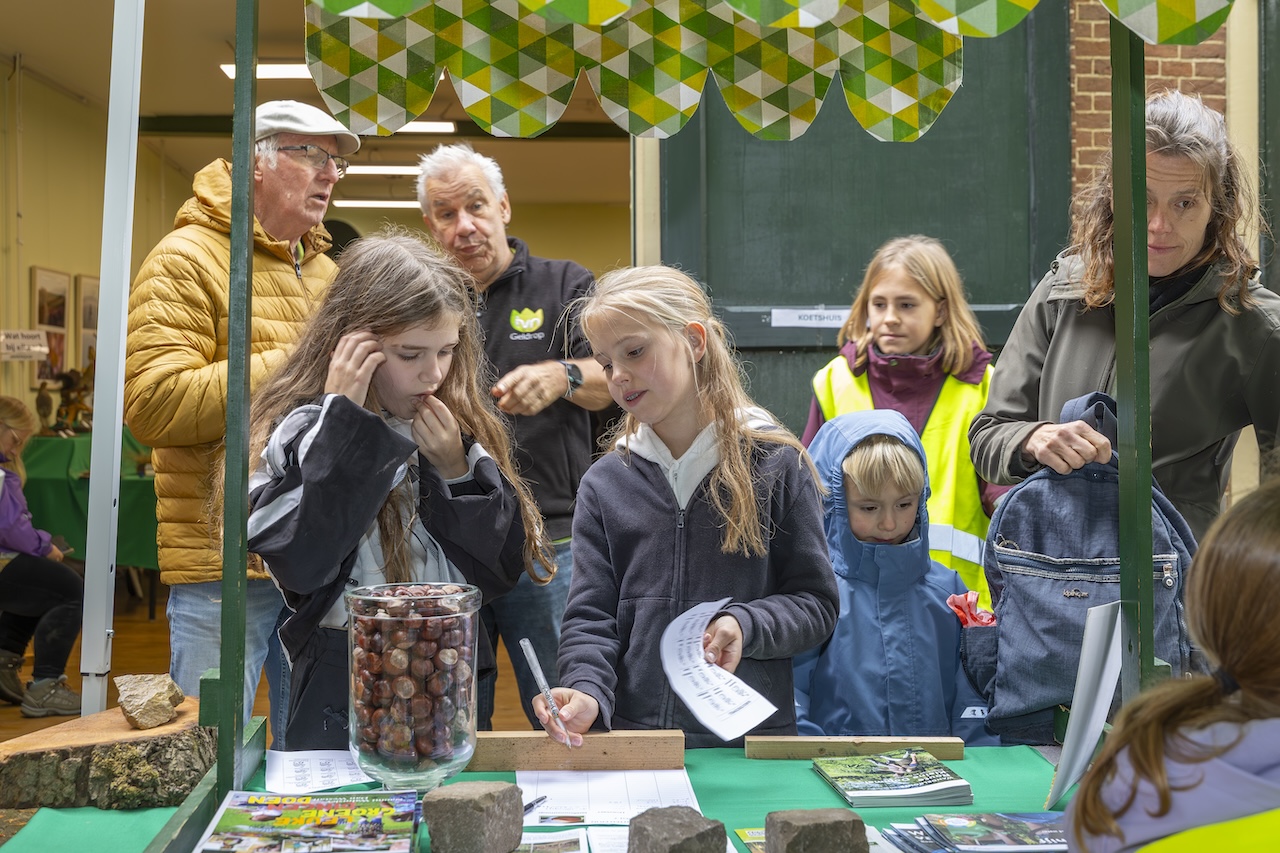Kinderen bij een kraam met folders en kastanjes, omringd door volwassenen.